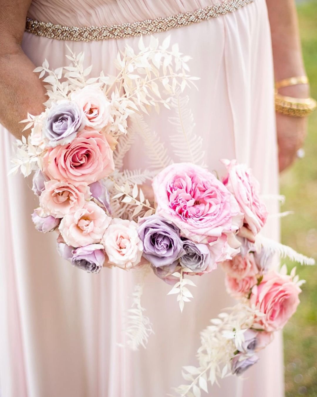 A bride elegantly holds a Hogarth Curve Bouquet filled with vibrant pink and purple flowers, showcasing her radiant beauty.